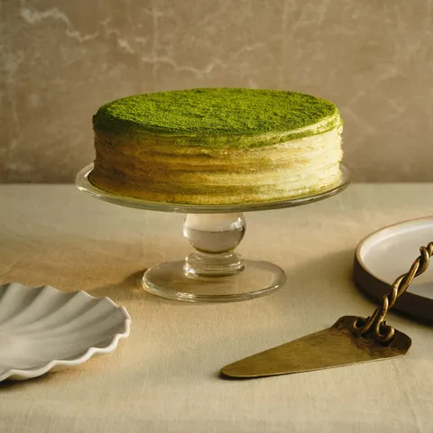A Lady M Green Tea Mille Crêpes on top a clear cake stand set on top a beige linen.