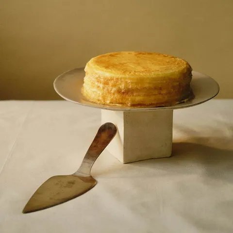 A whole 6-inch Lady M Signature Mille Crêpes on top a cake stand set over a white linen.