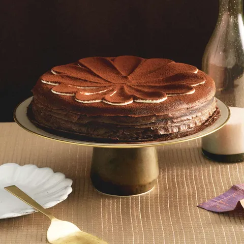 A whole Lady M Tiramisu Mille Crêpes on top a cake stand set over a beige linen.