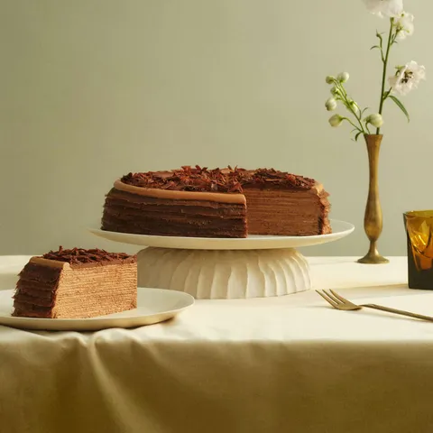 A Lady M Amedei Chocolate Mille Crêpes with one slice served on a plate over a yellow linen.