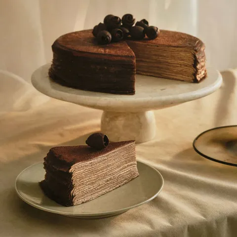 A Lady M Chocolate Mille Crêpes with one slice served on a plate, set on top a white linen.