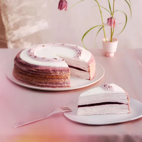 A Lady M Lavender Mille Crêpes with one slice served on a plate, set on a light purple linen.
