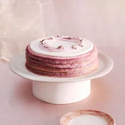 A whole Lady M Lavender Mille Crêpes on top a white cake stand, set over a light-purple linen.