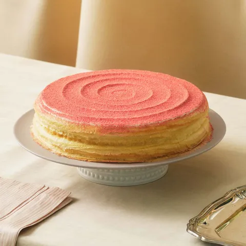 A Lady M Strawberry Swirl Mille Crêpes on top a white cake stand, set on top a linen table cloth.