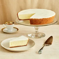 A Lady M Gâteau Nuage on top a clear cake stand with one slice served on a plate over a beige linen.