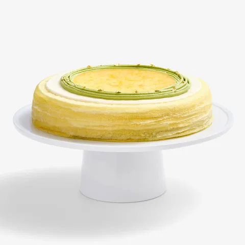 A whole Lady M's Yuzu Matcha Mille Crêpes sitting on top of a white round cake stand.