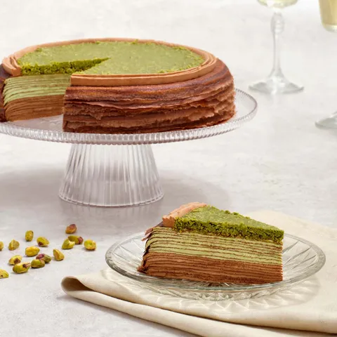 Lady M's Dubai Chocolate Mille Crêpes and slice with green layers, on glass stand with pistachios.