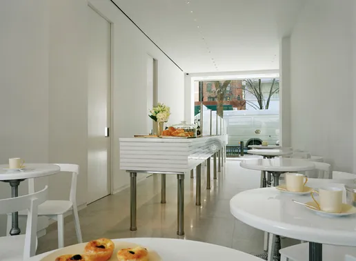 Lady M Upper East Side minimalist white interior with ribbed service counter and small round tables. White chairs and teacups contrast with light flooring, while pastries and cakes are displayed in foreground. Large windows show 78th Street view with brick buildings and trees.