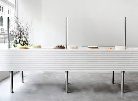 Lady M Bryant Park minimalist cake display: white ribbed counter, chrome, cakes, white flowers/branches. Concrete floor, bright.