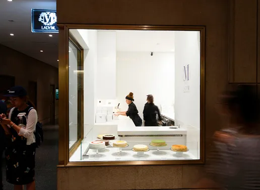 Lady M Rockefeller Center Boutique storefront with illuminated logo sign. Display window showcases various cakes and crepes on white pedestal cake stands. Staff in black uniforms work inside the bright white interior and cake boxes with the 'M' logo are visible on shelves behind the counter.