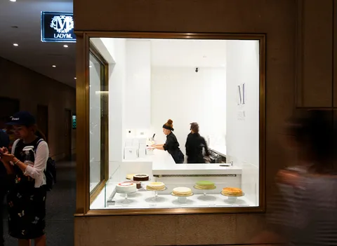 Lady M Rockefeller Center storefront: lit logo, cakes/crepes on white stands, staff in black, 'M' logo boxes. Bright.