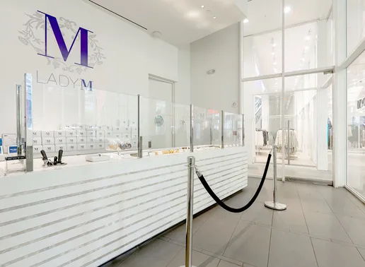 Bright, modern interior of Lady M World Trade Center Boutique featuring a long white cake display with horizontal marble detail and clear glass partitions. A large purple "M" logo with floral accents and "LADY M" in smaller letters is on the white wall above the cakes. The floor is light gray tile, and glimpses of a brightly lit storefront are visible in the background.