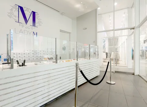 Bright, modern Lady M WTC: long white marble cake display, purple floral "M" logo, light gray tile. Storefront glimpse.