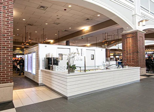 A brightly lit Lady M Cake Boutique kiosk situated in the Woodbury Common Food Court. The kiosk has a white cake display with glass showcasing cakes, and a white backdrop with the purple "LADYM" logo.