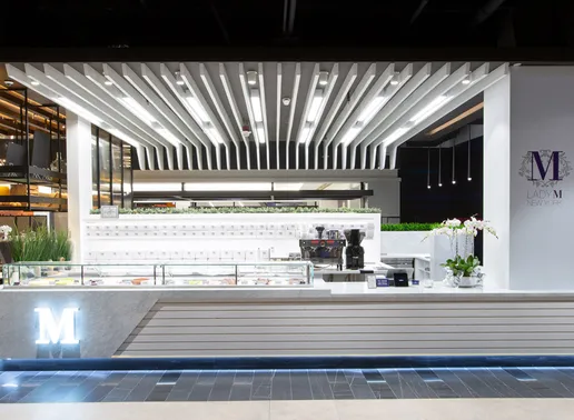 Lady M's modern and stylish dessert kiosk at American Dream featuring a sleek white design with clean lines and slatted paneling. The counter showcases an illuminated "M" logo, and a glass display case is filled with an assortment of colorful cakes. Overhead, geometric ceiling panels with integrated lighting add a contemporary touch, while fresh greenery and orchids enhance the inviting atmosphere.