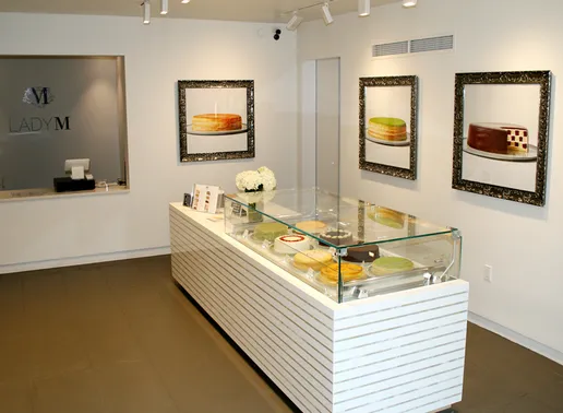 Interior of a Lady M Boston featuring a white display counter with glass case containing cakes. On the walls are framed photographs of signature cakes, and a Lady M logo is visible on the left wall.