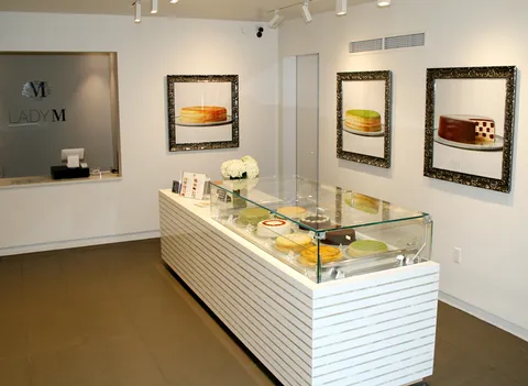 Lady M Boston interior: white display counter, cakes, framed cake photos, Lady M logo on wall.