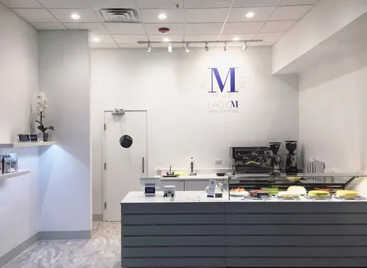 Lady M Chicago boutique with white walls featuring the Lady M logo. The space has a modern gray counter with a glass display case showing various cakes. A coffee machine sits on the counter, and a white orchid decorates the space.