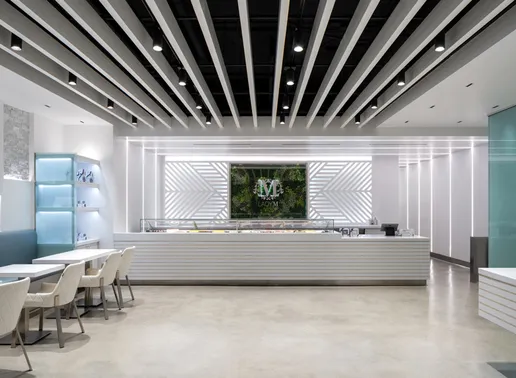 Interior of Lady M Sawtelle featuring a long, white cake display. Behind the counter is a floral "M" logo with greenery. Ceiling features black and white parallel beams. Small white tables and light-colored chairs are arranged on the left side of the space.