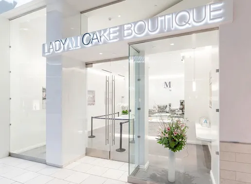 Storefront of LADY M CAKE BOUTIQUE in Arcadia, CA within The Shops at Santa Anita featuring illuminated signage, glass doors, bright white interior, and a floral arrangement by the entrance. The design is modern and elegant with minimalist aesthetic.