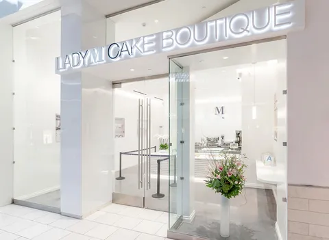 LADY M Arcadia at Santa Anita storefront: illuminated sign, glass doors, bright white interior, floral arrangement. Modern, elegant.