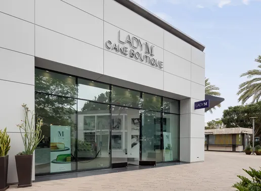 Modern white Lady M San Diego storefront with large glass windows and silver signage. The entrance features glass doors and display windows showing "M" branding inside. Decorative plants are positioned near the entrance, with palm trees visible to the right. The contemporary design sits on a paved walkway.