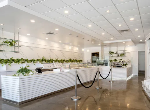 The minimalist white interior of Lady M's Los Altos boutique with horizontal slat counters, black queue barriers, display cases with colorful cakes, and decorative plants. Features bright recessed lighting and polished concrete floors, creating a clean, modern atmosphere.