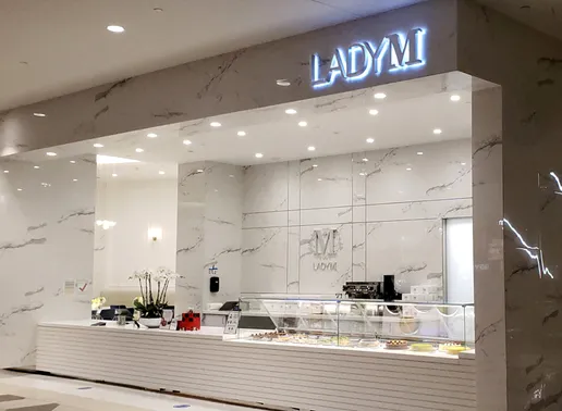 An elegant Lady M counter located at Westfield Valley Fair with a sleek design featuring white marble walls and surfaces. The illuminated "LADY M" sign glows above the service area. The display case showcases colorful cakes, while a small white orchid decorates the counter. The minimalist space is brightly lit with recessed lighting.