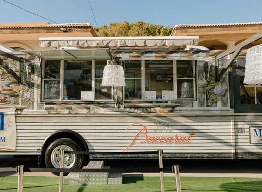 A luxury cake truck with "Baccarat" and "LADY M" logos on its side. The silver trailer features a retractable awning, serving windows with pendant lamps, Baccarat chandeliers and is parked on a green surface.