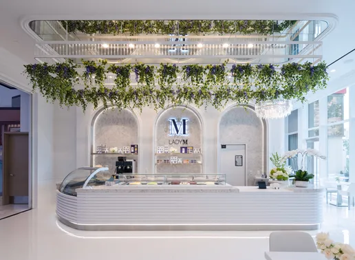 Interior of Lady M at Resorts World Las Vegas featuring an elegant white cake display counter with glass display case. Behind the counter are three arched alcoves with marble backing and the Lady M logo. Hanging greenery and plants create a garden-like canopy above the counter, and a crystal chandelier adds a touch of luxury to the bright, airy space.