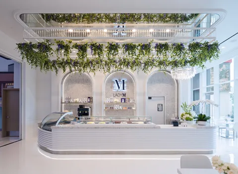 Lady M Resorts World LV interior: white cake counter, glass display, marble alcoves, logo, hanging greenery, crystal chandelier.