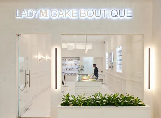 Front entrance of Lady M Cake Boutique at The Galleria in Houston with illuminated signage, a modern white interior, display case with cakes, and a person behind the counter.