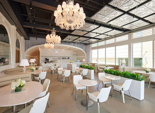 Elegant interior of Lady M Houston Westchase boutique featuring crystal chandeliers, bright natural lighting through glass windows, white round tables with comfortable seating, and an arched service counter in the back displaying cakes, ice cream, and coffee.