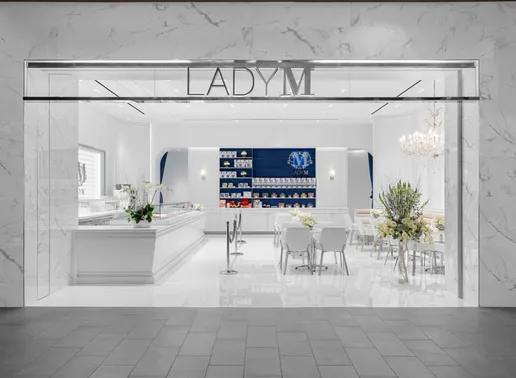 Elegant storefront of Lady M at Tysons Galleria featuring a marble exterior with the LADY M logo prominently displayed above the entrance. Inside, a pristine white interior showcases a cake display counter on the left, a blue accent wall with product displays in the back, and several small white tables and chairs for guests to enjoy their desserts.