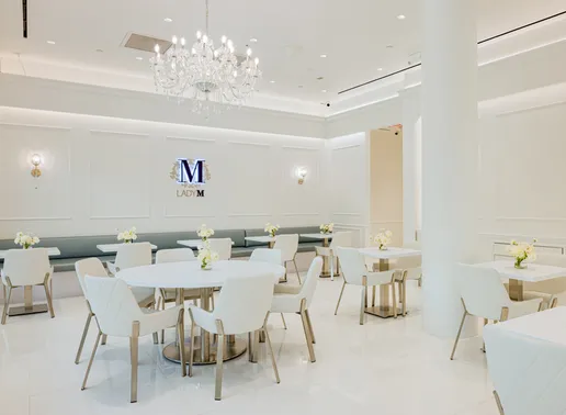 Elegant dining area of the Lady M Bellevue boutique featuring a luxurious crystal chandelier, pristine white walls with decorative paneling, and the Lady M logo displayed prominently. The space includes white tables with white quilted chairs, fresh flower arrangements, and a soft gray banquette seating along the wall, all creating a sophisticated atmosphere for guests.