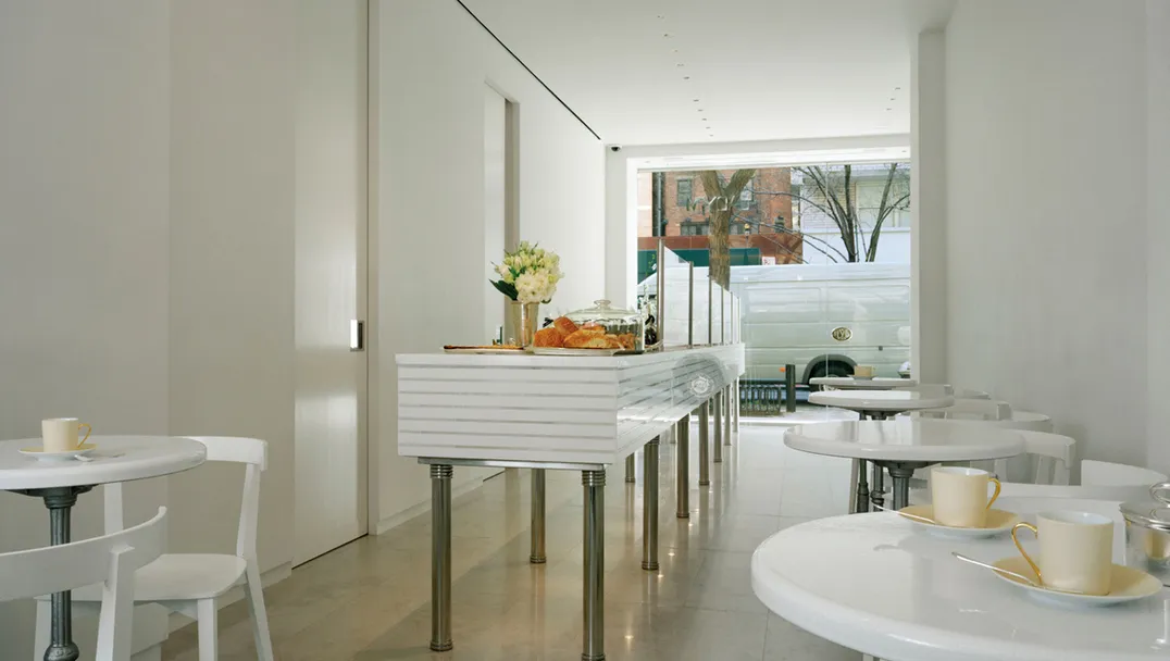 Lady M Upper East Side minimalist white interior with ribbed service counter and small round tables. White chairs and teacups contrast with light flooring, while pastries and cakes are displayed in foreground. Large windows show 78th Street view with brick buildings and trees.