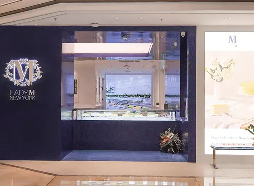The Lady M Grand Gateway 66 Shanghai Boutique storefront with a navy blue exterior featuring an illuminated Lady M logo. A display window shows the cake showcase inside the minimalist white interior, with a digital board visible on the right side.