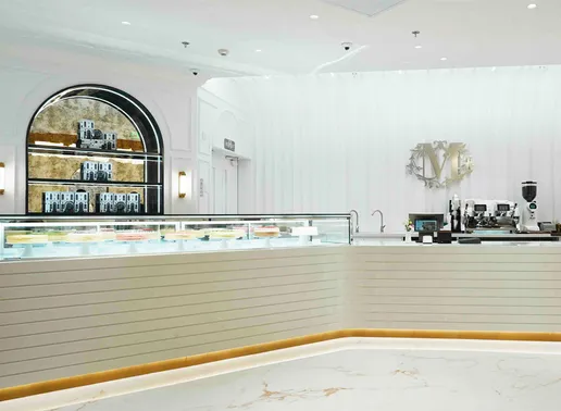 The elegant interior of Lady M Shanghai IFC Boutique featuring a white counter with a glass display case showcasing cakes. The space includes an arched display shelf with candy boxes, a marble floor with gold accents, and the signature Lady M logo mounted on the white wall beside the coffee station.
