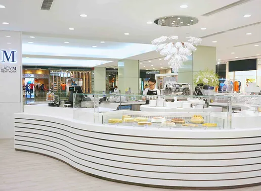 Lady M Tsim Sha Tsui Hong Kong Boutique with a stylish curved white counter featuring horizontal stripes. It includes glass display cases showing cakes, and is staffed by an employee in a black apron. The space is elegantly lit with a crystal chandelier and features the blue Lady M logo on the left wall.