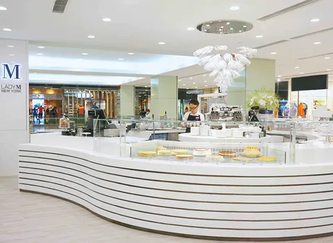 Lady M Tsim Sha Tsui HK: curved white striped counter, cake displays, staff in black apron, crystal chandelier, blue logo.