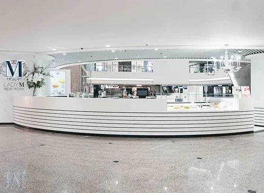 Lady M Causeway Bay Times Square Hong Kong with a white horizontal-striped counter and glass display cases. The boutique features the blue Lady M logo with decorative floral elements on the left wall, a crystal chandelier, and white floral arrangements complementing the sleek, minimalist design.