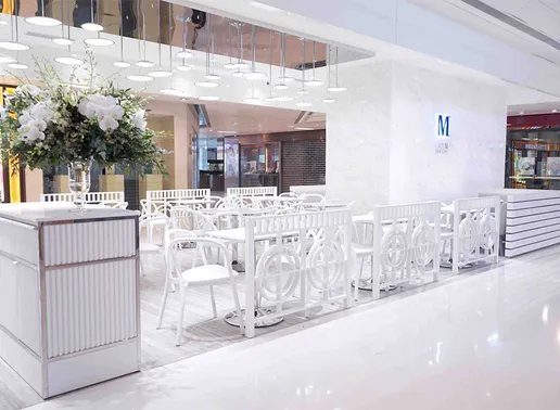 Lady M Sha Tin New Town Plaza Hong Kong Boutique dining area featuring elegant white furniture with decorative railings, marble walls with the blue Lady M logo, and modern pendant lighting. White tables and chairs are arranged in the open space, with a large floral arrangement visible on a striped pedestal in the foreground.