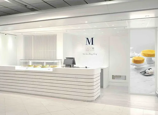 Lady M Hong Kong International Airport Boutique featuring a white counter with horizontal striped paneling and a small display case showing cakes. The clean interior includes the Lady M logo on the back white wall, a point-of-sale system, and a large digital display of cakes on the right side. The space embodies the brand's elegant, contemporary aesthetic.