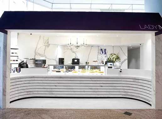 Lady M Quarry Bay Taikoo Place Hong Kong Boutique kiosk with a navy blue awning and white LADY M lettering. The elegant marble-walled interior features the signature horizontal striped counter with glass display case showing colorful cakes. The blue Lady M logo appears on the back wall, and a crystal chandelier adds a touch of luxury to the minimalist design.