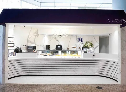 Lady M Quarry Bay HK kiosk: navy awning, white "LADY M", marble walls, striped counter, colorful cakes, blue logo, crystal light.