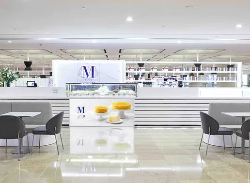 Lady M Admiralty Pacific Place Hong Kong Boutique with a white counter featuring cake imagery and horizontal stripes. The space has the blue Lady M logo on the back wall, display cases, gray dining tables with chairs, and shelving for merchandise. Floral arrangements add elegant touches.