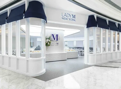 Lady M Macau Londoner Boutique storefront with white framing, navy blue awnings, and large glass windows. The entrance displays the "LADY M NEW YORK" sign above, while inside shows the signature white striped counter, blue Lady M logo on the wall, and floral arrangements in the bright, elegant space.