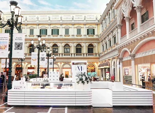 Lady M Macau Venetian Boutique kiosk with signature white striped counters in a European-style indoor shopping center featuring Venetian architecture. The boutique displays the blue Lady M logo and glass cases with cakes against a backdrop of ornate buildings with columns, arches, and decorative facades.