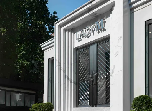 Exterior view of Lady M's Taipei Flagship Boutique with a modern architectural design featuring a white marble facade with vertical linear elements. The entrance has striking geometric patterned glass doors with an "M" handle design, and the silver LADY M logo is prominently displayed above. Small manicured shrubs frame the entrance against a bright blue sky.