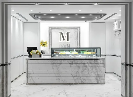 Lady M Regent Taipei Boutique with a marble counter and glass display case showing various cakes. The elegant space features a framed Lady M logo on the marble back wall, recessed ceiling lighting, and yellow floral arrangements. The minimalist white and gray color scheme creates a sophisticated atmosphere.